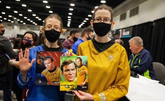 Two Star Trek cosplayers pose with their signed photo of William Shatner. Shatner is seen in the background.