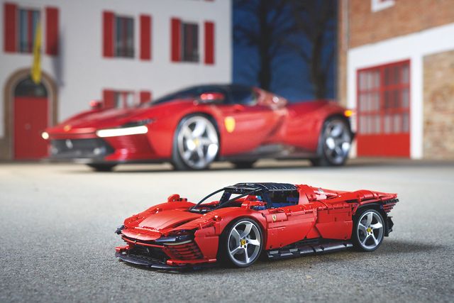 Two blurred buildings in the background; one white with red trimmings and doors, and a brown one with red trimming and red doors. There is a red Ferrari Daytona parked in the background. In the foreground and in focus, is a Lego version of the car behind it.