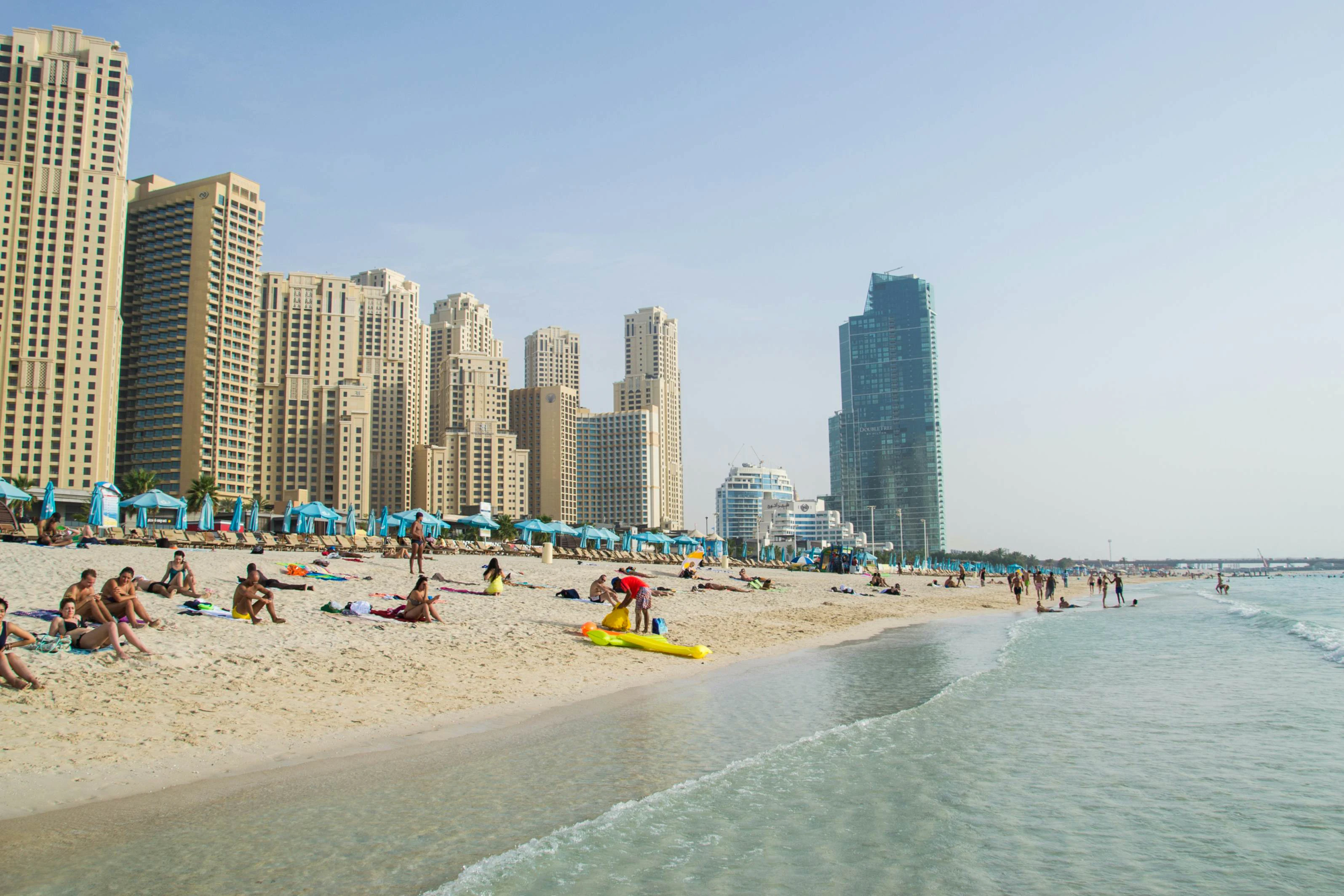 Beachfront scene with people sunbathing and enjoying the water, with tall buildings and blue umbrellas in the background.