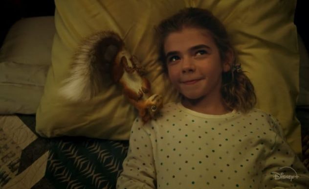 A girl with a white dotted PJ top lays in bed smiling next to a CGI squirrel