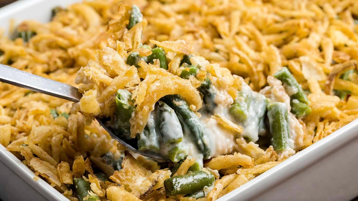 A spoon dips into a green bean casserole to show a spoonful of green beans, butter and fried onions.