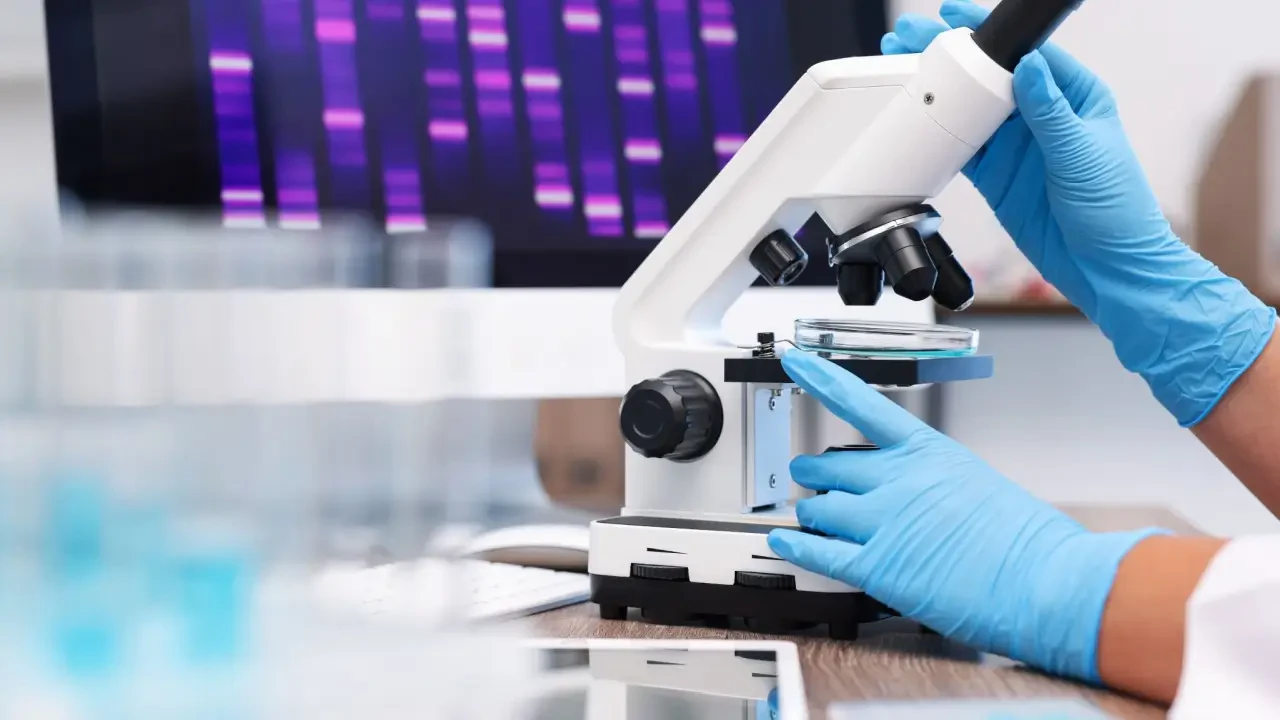 Microscópio sendo operado por profissional com luvas em laboratório, analisando amostra em placa de Petri, com tela de sequenciamento genético desfocada ao fundo — conceito de pesquisa científica, biotecnologia e diagnóstico laboratorial.