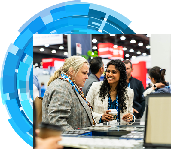 Two women engaged in conversation at a bustling trade show, surrounded by various booths and attendees
