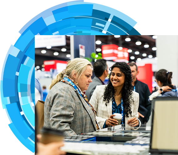 Two women engaged in conversation at a bustling trade show, surrounded by various booths and attendees