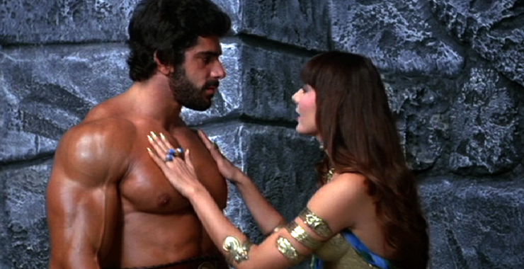 Lou Ferrigno is standing next to a brunette lady. She has her hands on his chest (he has no shirt on). She has a lot of rings on her finger and the gold bands on her arms. She is wearing gold and blue armor. Behind them is a stone wall.