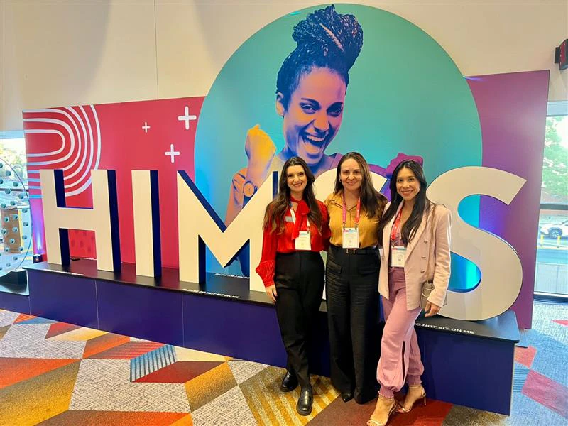 Três mulheres posam sorrindo em frente a um grande painel colorido com a sigla “HIMSS”. Elas usam crachás de evento e roupas formais — uma de blusa vermelha e calça preta, outra de blusa mostarda com calça preta, e a terceira com blazer rosa e calça lilás. Ao fundo, há um design vibrante com uma ilustração feminina e elementos gráficos modernos, em um ambiente interno de evento com carpete colorido.
