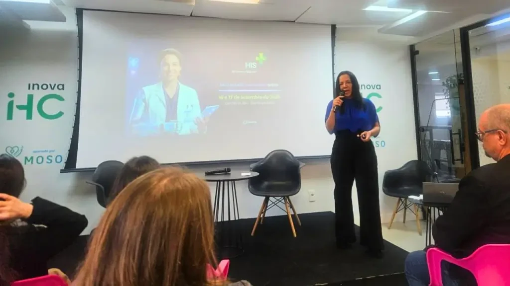 Mulher palestrando em palco com microfone durante o evento Healthcare Innovation Show (HIS). Ao fundo, projeção em tela com a marca "Inova HC" e informações sobre o evento de inovação em saúde. Em primeiro plano, público assiste à apresentação.