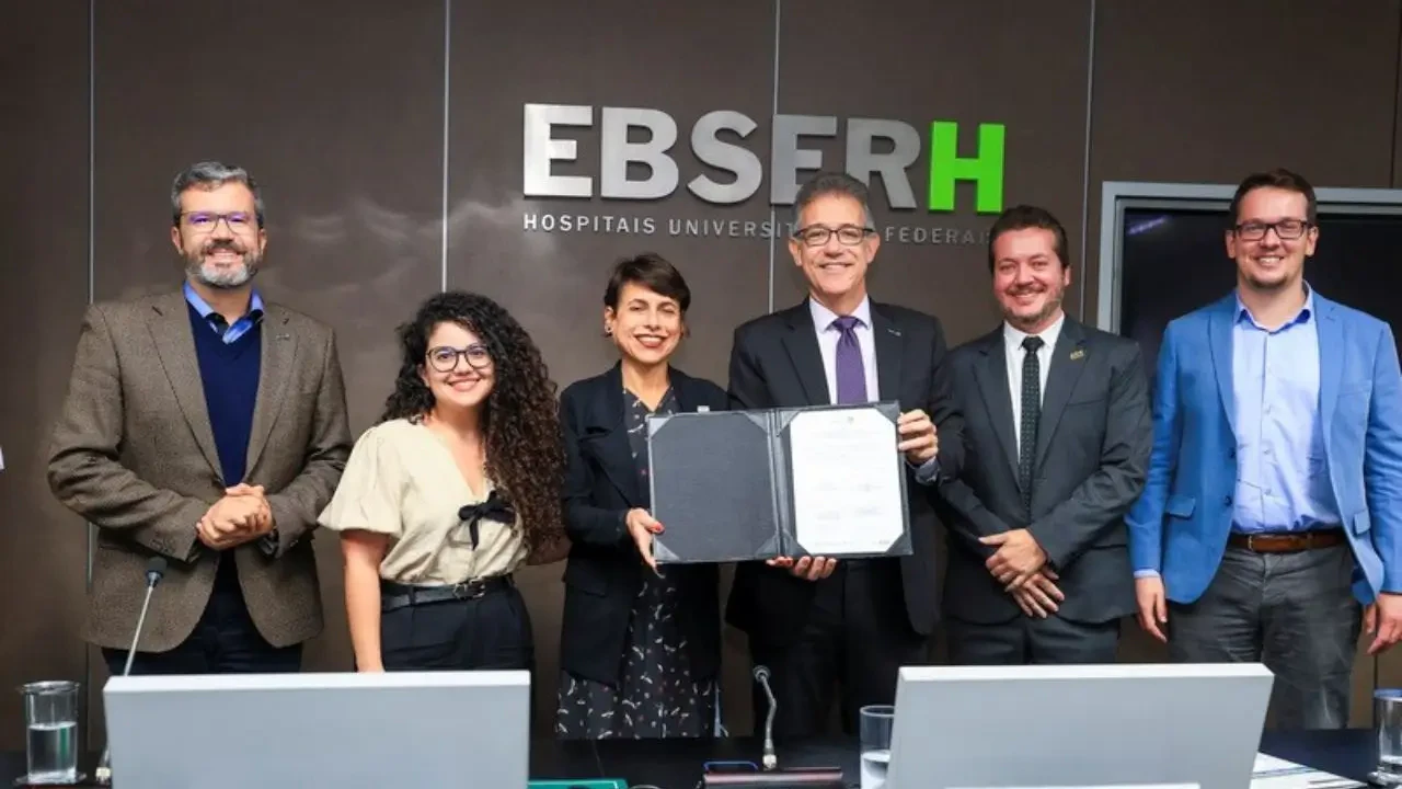 Grupo de executivos e autoridades posando para foto oficial durante cerimônia na EBSERH (Empresa Brasileira de Serviços Hospitalares). No centro, um representante segura um documento oficial assinado, simbolizando parcerias estratégicas no setor de hospitais universitários federais.