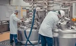 Dois técnicos operando reatores de aço inoxidável em um laboratório industrial de alta precisão, utilizando jalecos brancos, redes de cabelo e EPIs em ambiente controlado.