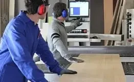 Vista de um centro de usinagem CNC de madeira moderna, onde dois marceneiros com equipamentos de proteção auricular estão colaborando, um operando a tela de controle digital e o outro posicionando uma peça de madeira laminada para processamento de precisão, exemplificando a automação na marcenaria.