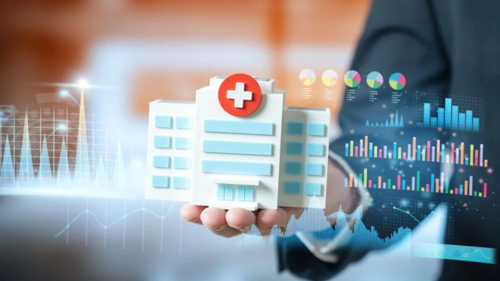 Mão segura maquete de hospital cercada por gráficos e indicadores digitais, representando gestão e infraestrutura hospitalar.