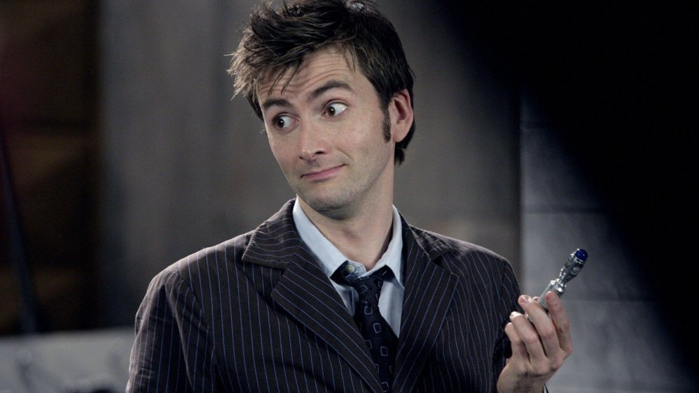 The Tenth Doctor from Doctor Who is wearing a striped suit with a blue shirt and paisley tie. He is holding up a small silver object in his hand and is shrugging at someone out of view.