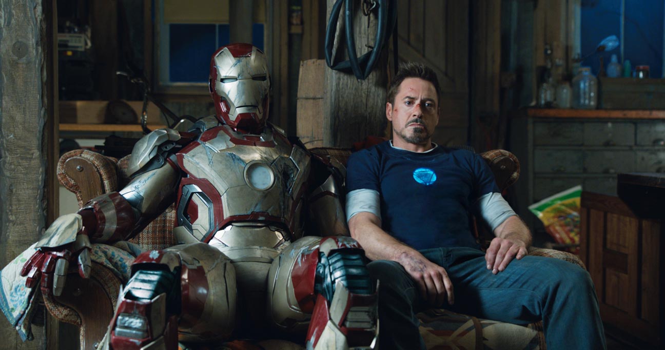 A roughed up Tony Stark sits next to a scratched up Iron Man suit on an old plaid couch