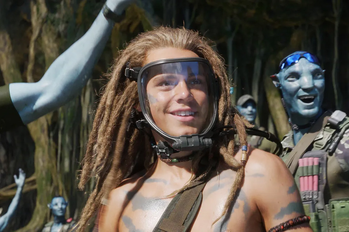 Spider from Way of Water smiles while wearing his mask & surrounded by Na'vi Avatars