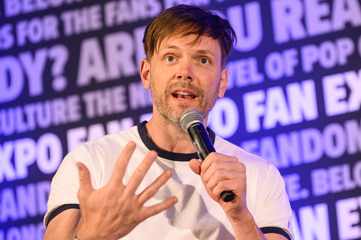 A headshot of Joel Mchale. He is holding a microphone and is in the middle of talking/asking a question. His brown hair is a bit tousled and he has some scruff. He is wearing a white t-shirt with a black collar. In the background, is the FAN EXPO background that repeats a variety of phrases.
