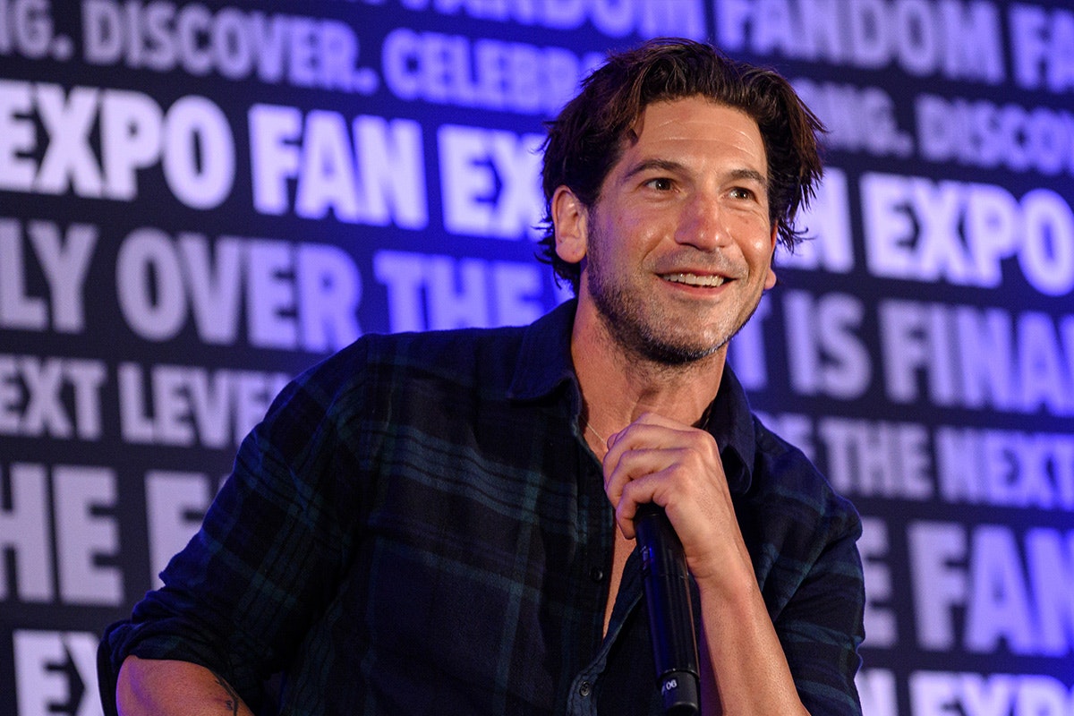 This is a headshot of Jon Bernthal holding a microphone. He is in the midst of answering a question so his mouth is open. His brown hair is tousled and her has a slight scruff. He is looking to the left of the image. He is wearing a blue button up flannel and is wearing 2 gold chains around his neck. In the background, is the FAN EXPO background that repeats a variety of phrases.