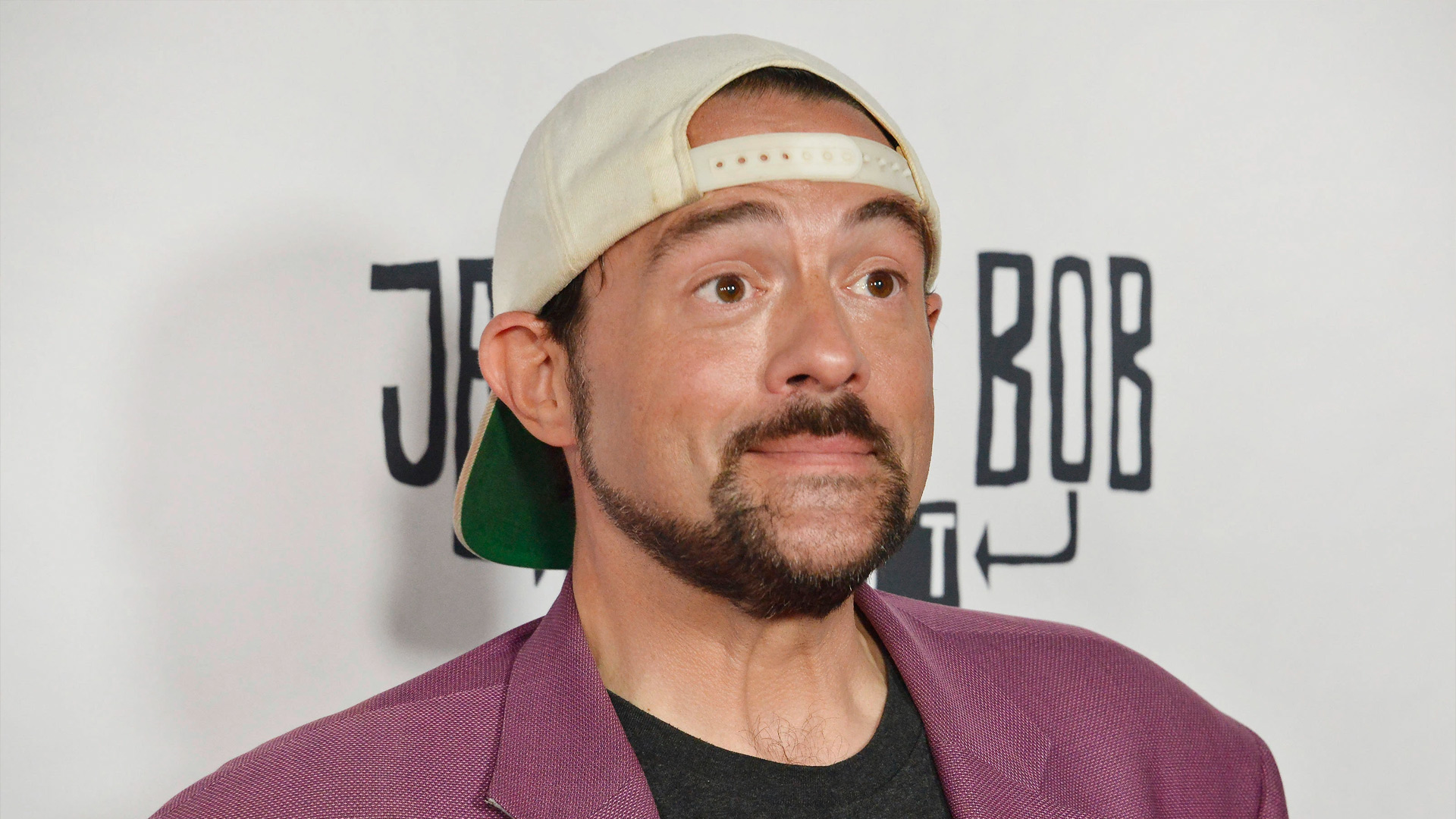 Kevin Smith smiling in a purple blazer with a white baseball hat on backwards