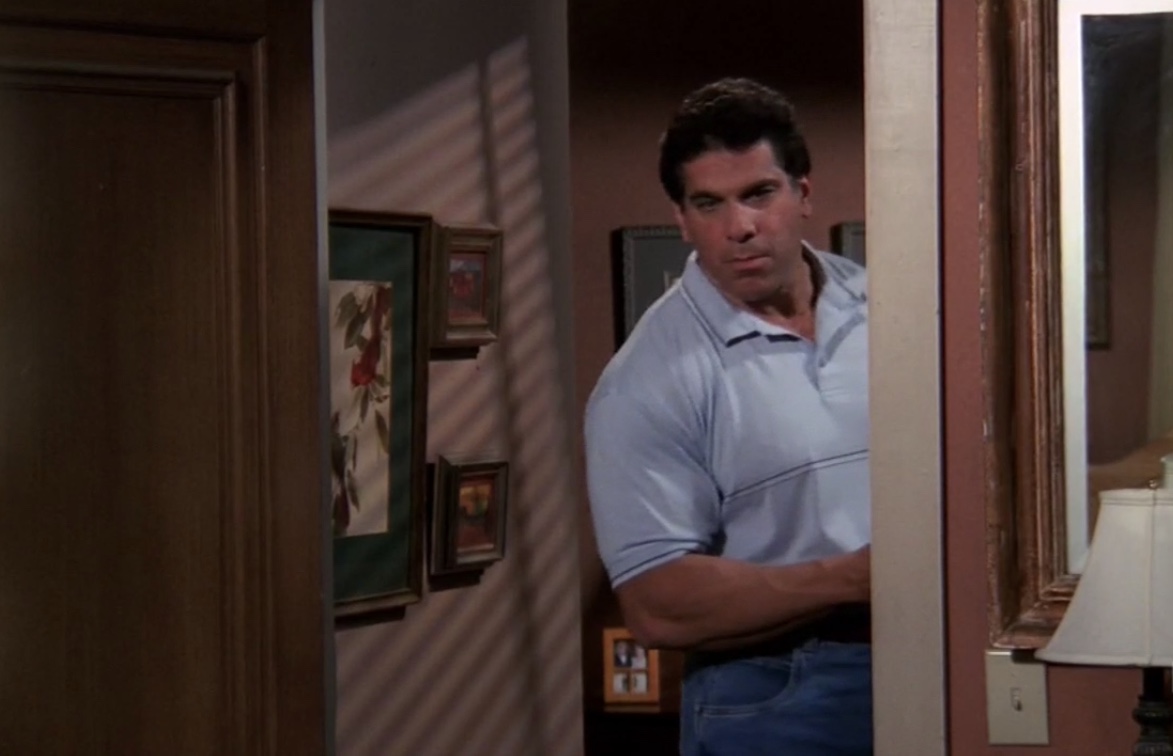 Lou Ferrigno is standing in a doorway peaking in. He has on a blue polo shirt and jeans. In the right side of the frame is a bit of a wall mirror and a lamp. Behind Lou and to the left is a wall full of pictures and photos.