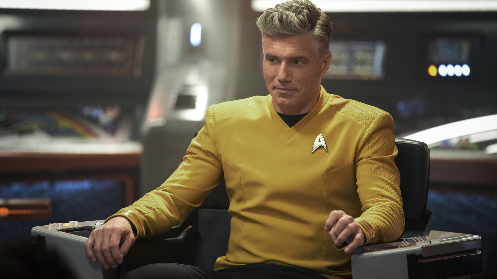 Christopher Pike sits in his captain's chair, wearing the signature yellow tunic. He smiles at the costar off screen