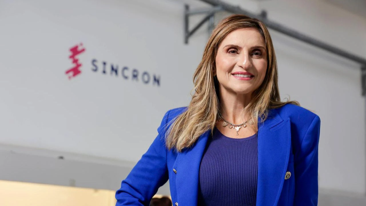 O Gemini disse Mulher sorridente de cabelos longos e loiros, vestindo um blazer azul vibrante, posa em frente a uma parede branca com o logotipo da empresa "SINCRON".