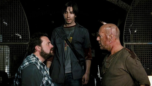 Kevin Smith opposite to Bruce Willis (bloodied from a fight). Behind them is Justin Long who is looking at Kevin. Behind them is a metal grate or fence.