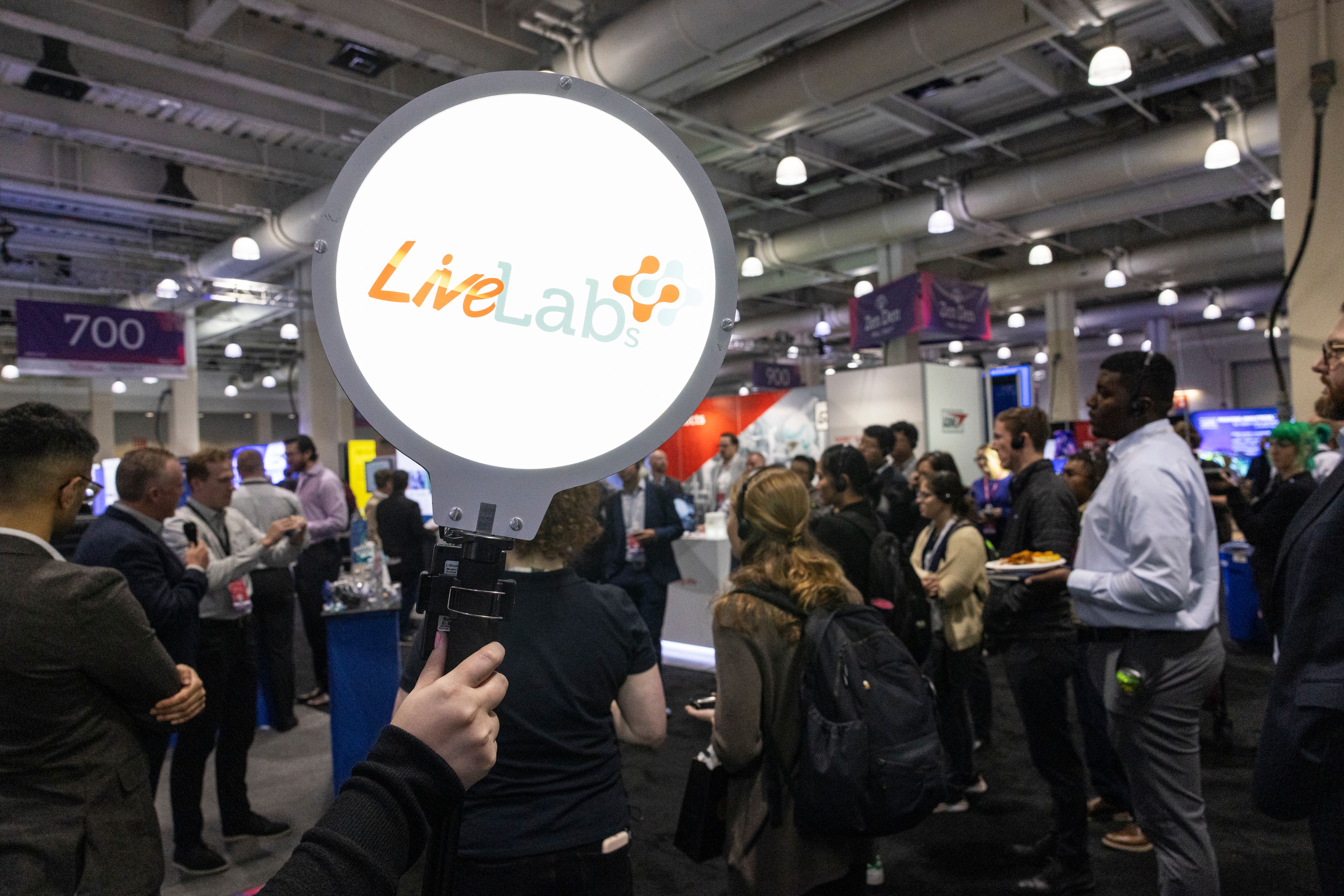 Arm holds up a Live Labs sign while a live demonstration takes place in the background with bioprocessing professionals at BioProcess International (BPI).
