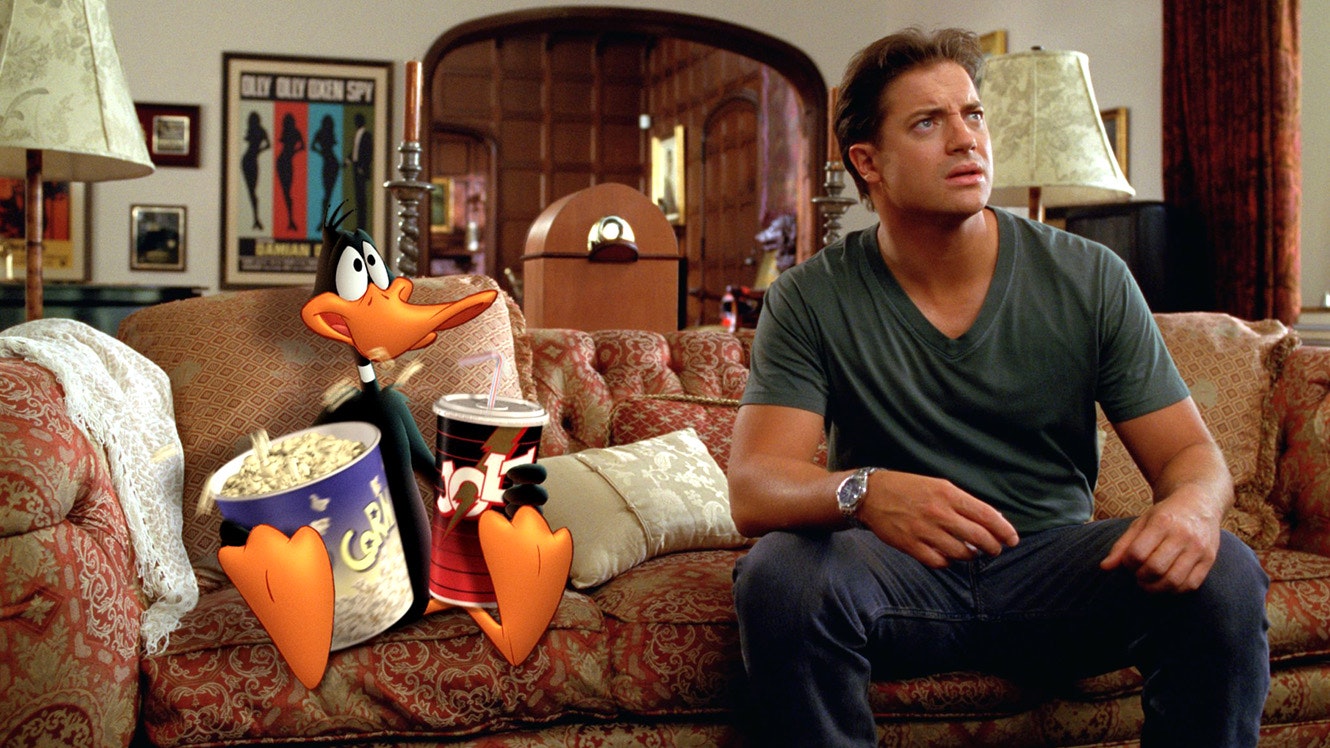 In a room on a couch, Daffy duck is on the left, holding popcorn and a big soda as he sits next to Brendan Fraser with a confused look on his face as he stares off screen