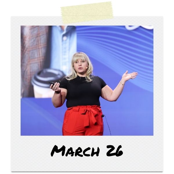 A photo of a woman speaking onstage in front of a blue background.