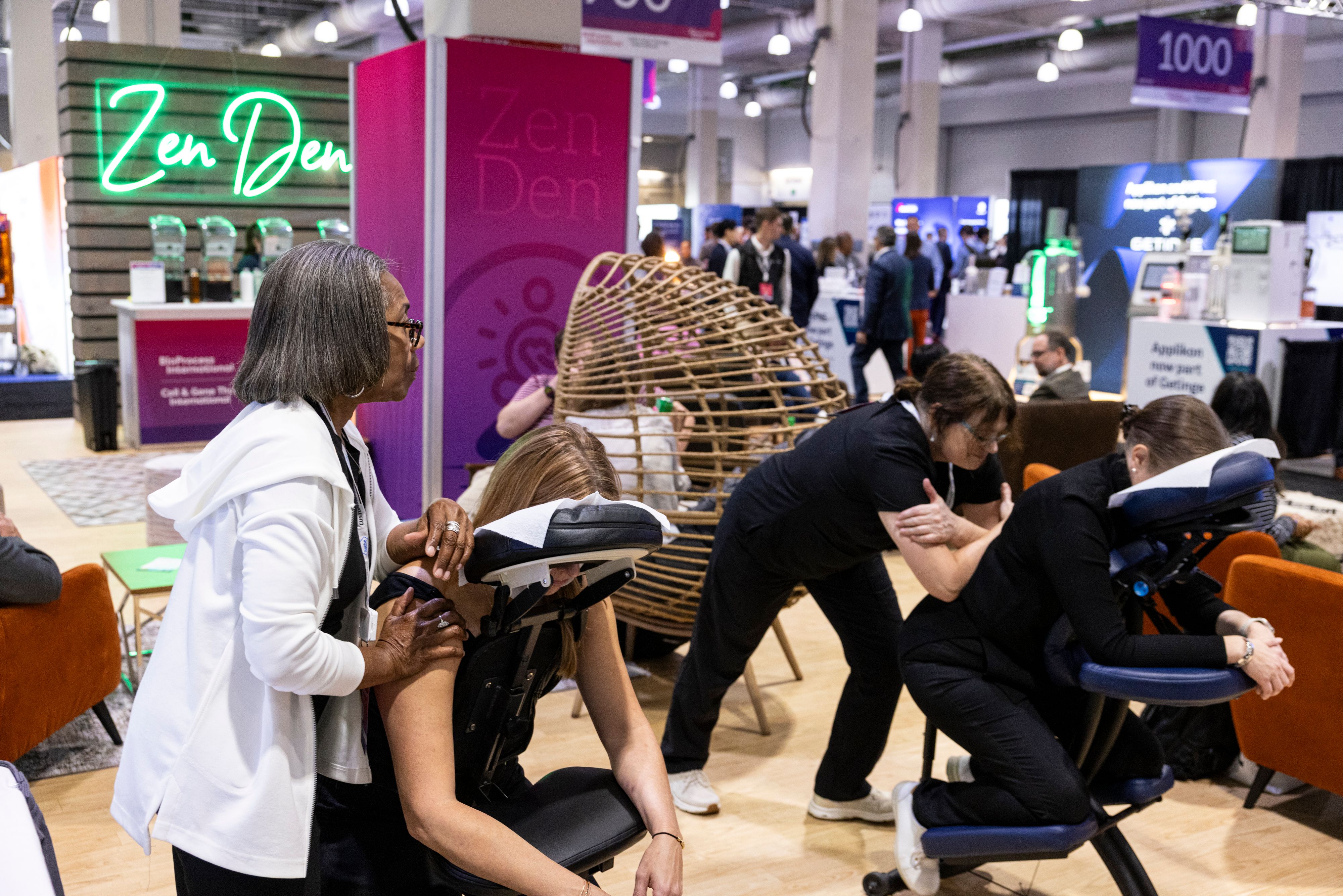Two BioTech Week Boston biopharma attendees receive massages in massage chairs with masseuses pressing on their backs. Zen Den signs are in the background.