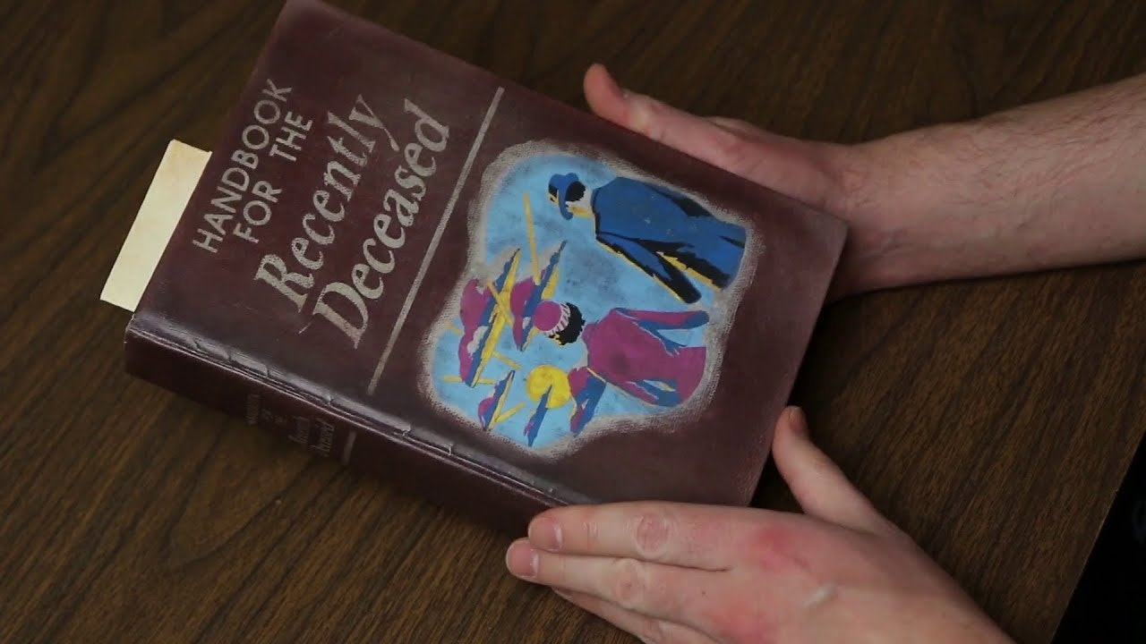Two hands hold a book marked copy for the Handbook for the Recently Deceased on a dark wood table