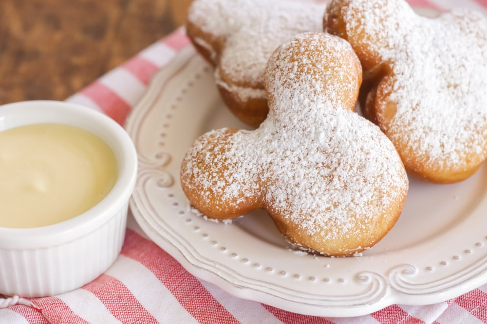 three Mickey Mouse head shaped beignets are stacked on a white platter. There is a ramekin of vanilla dip next to the platter - both lay on a red and white striped table mat