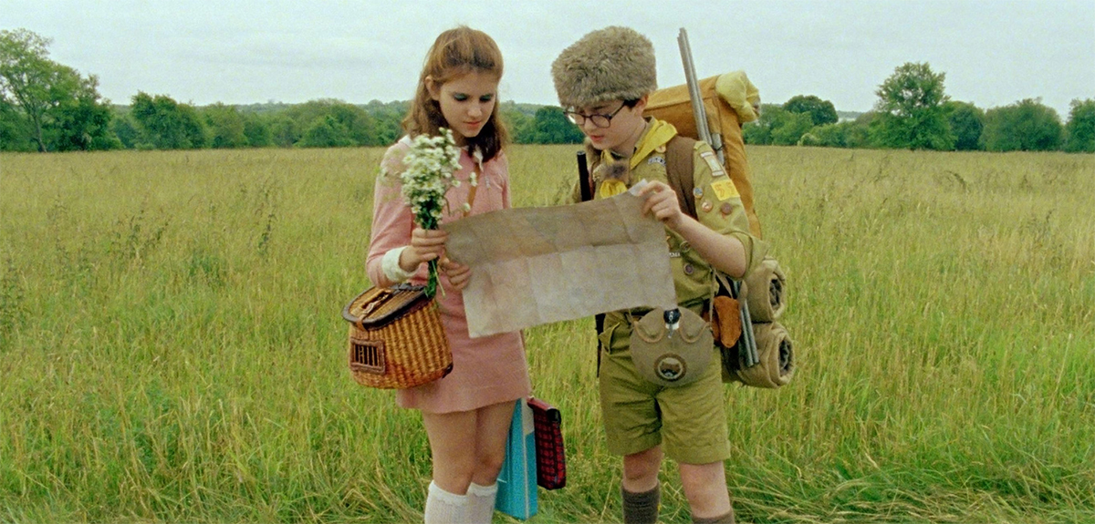 Suzy, with her bouquet of wildflowers, and Sam, in his scouts suit, hold a map together as they look for their destination in a field.