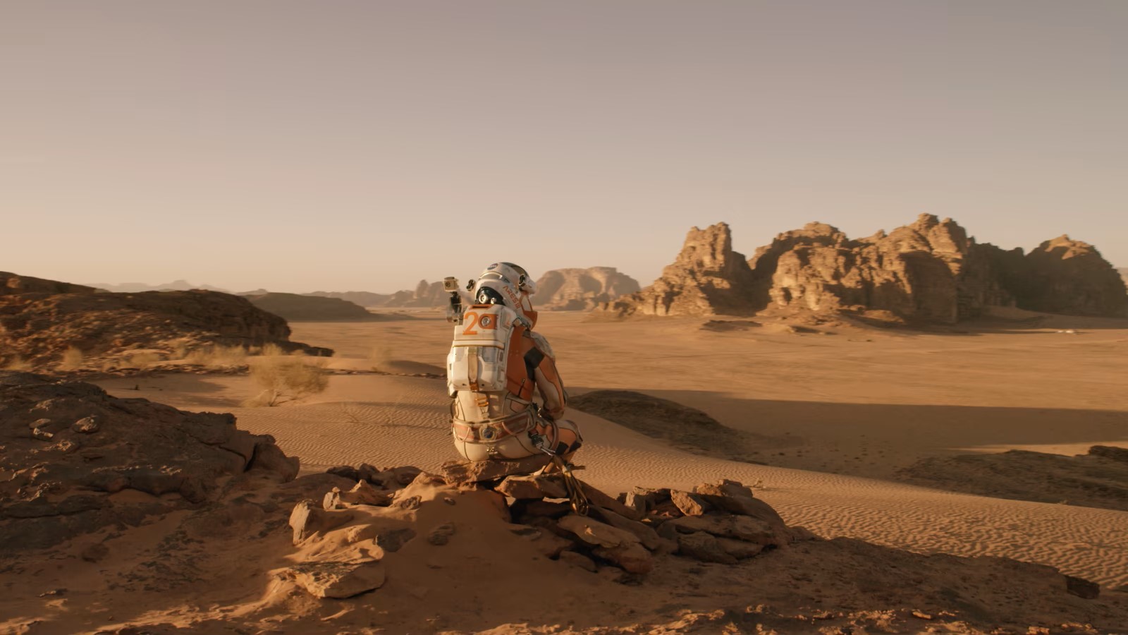 The Martian sits on a rock in the middle of the desert and looks off into the distance.