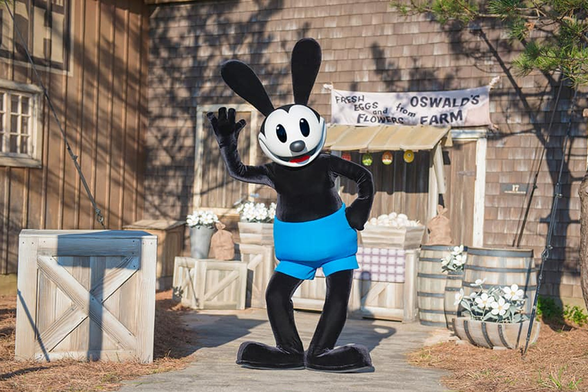 A mascot version of Oswald Rabbit waves hello posing in front of a barn that says "Oswald's Farm"