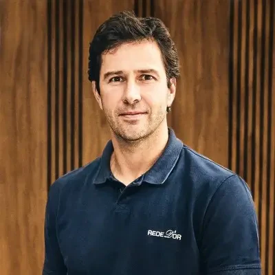 Paulo Moll, CEO da Rede D'Or, vestindo camisa polo azul-marinho com o logotipo da Rede D'Or, posando para foto contra fundo de painel de madeira.