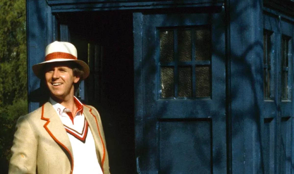The Doctor stands in the doorway to the TARDIS, smiling as he stares off to his companion off screen