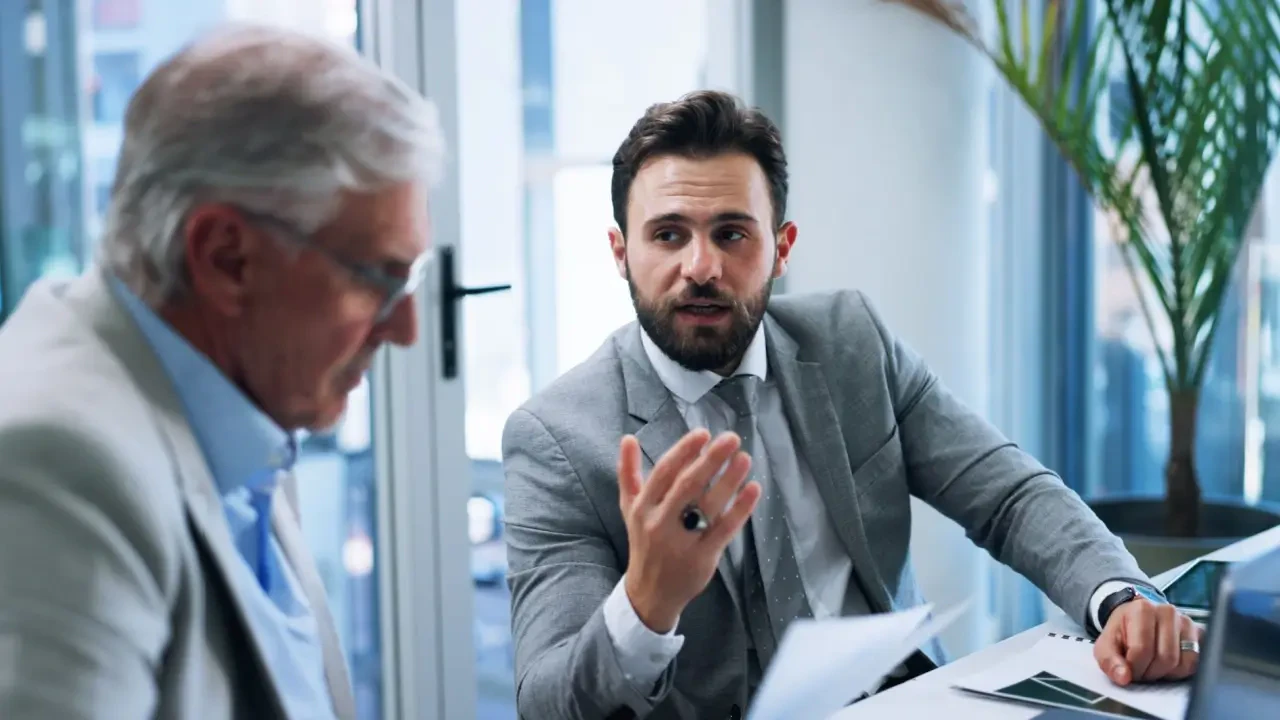 Gestor de terno cinza gesticulando durante reunião estratégica com colega sênior, exemplificando clareza na comunicação e direcionamento de equipe em ambiente corporativo.