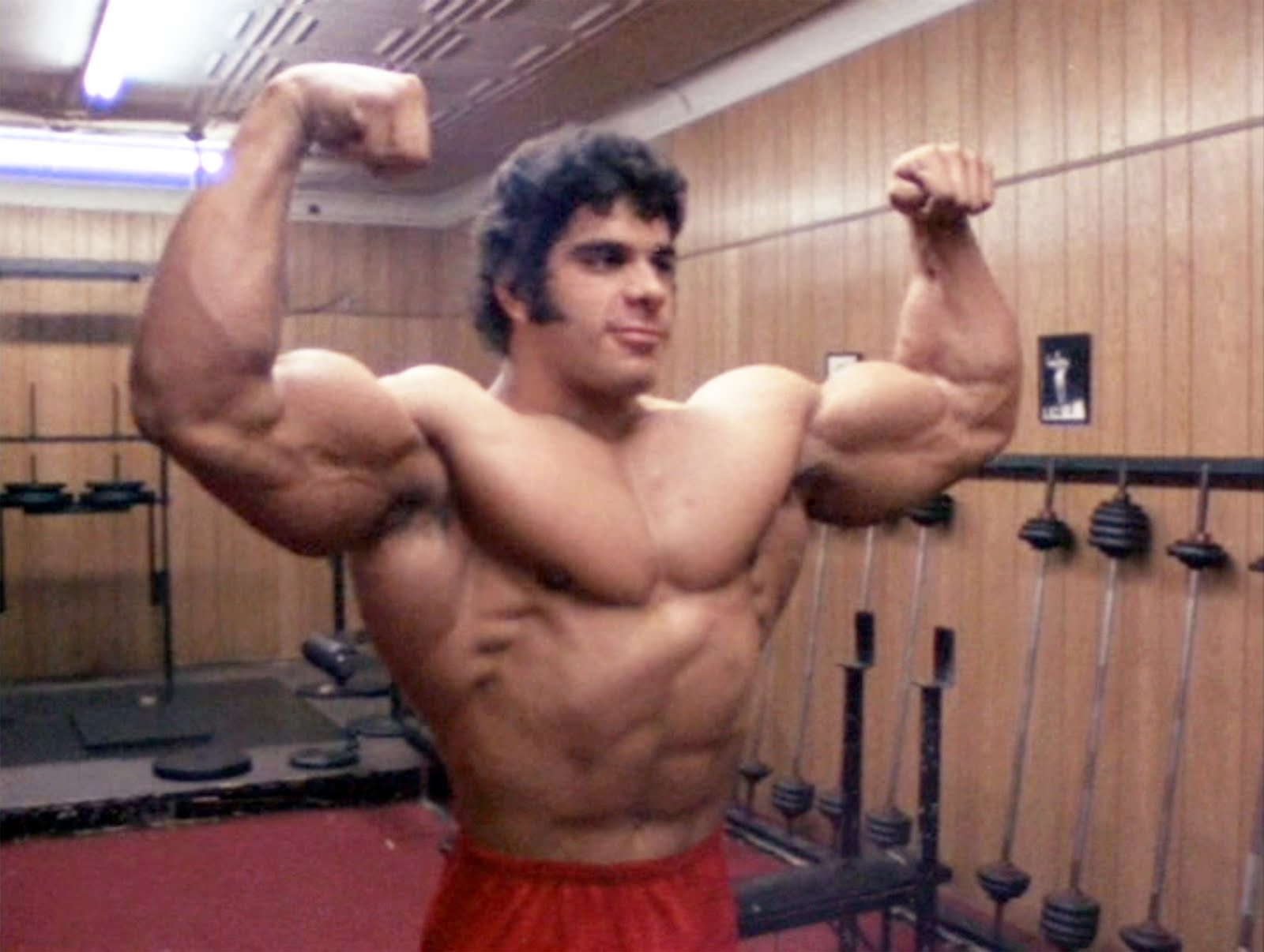 Lou Ferrigno is in a gym with wooden walls and various weights around him. He is raising his arms in a flexing position. He is shirtless and wearing red gym shorts.