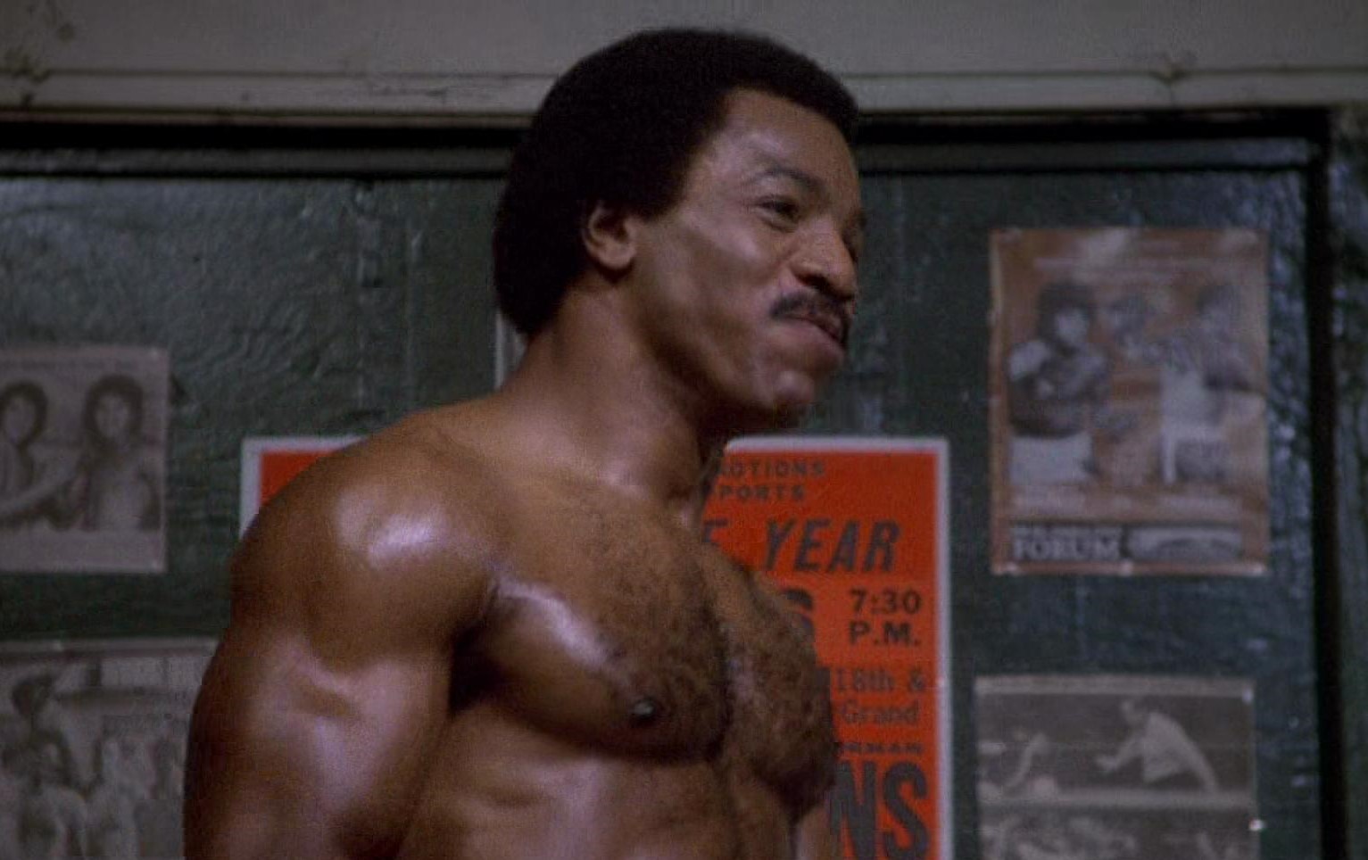 Shirtless Carl Weather smiling in front of a bunch of photos/posters