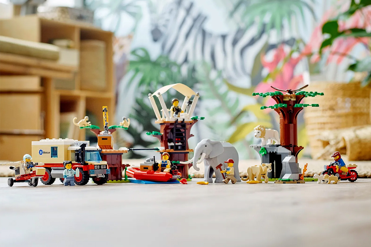 A Lego truck next to a Lego girl helping an Elephant. In the bottom right, a Lego man is trying to put out a wildfire. In the back is 3 trees with an assortment of African animals and Lego people interacting with each other.