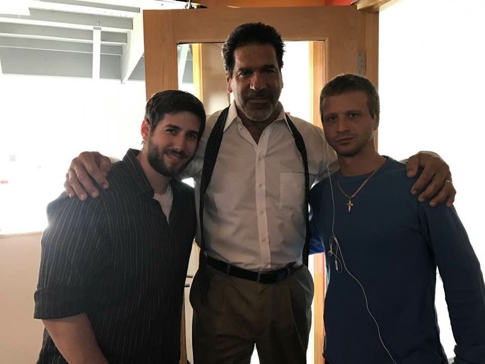In a bare room and an open door, Lou Ferrigno is in the center in a white shirt, brown pants and suspenders as he has his arms around two guys. The one on the left is wearing a button up stripped shirt and the one on the right is wearng a blue shirt, a cross necklace and earbuds in one ear.