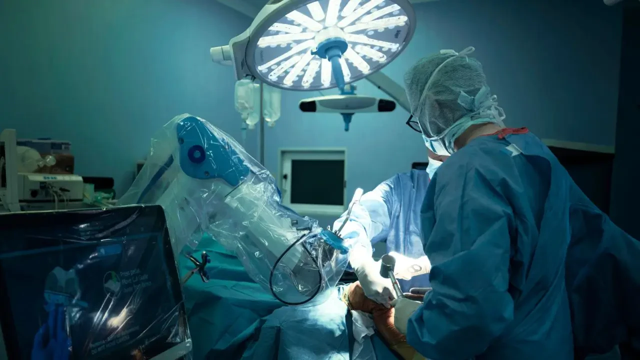 Foto de uma cirurgia robótica em andamento em um centro cirúrgico moderno. Um cirurgião com paramentação azul opera o paciente auxiliado por um braço robótico de alta tecnologia revestido por plástico protetor. Imagem ilustra o avanço da cirurgia minimamente invasiva, tema central na Hospitalar 2026.