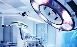 Foco cirúrgico de LED moderno em primeiro plano com braços articulados e monitores de sinais vitais ao fundo, compondo uma sala de operação equipada com tecnologia de ponta em ambiente hospitalar estéril.