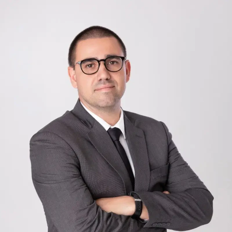 Saulo Lemos de braços cruzados, usando terno cinza escuro, camisa branca, gravata preta e óculos de armação preta.