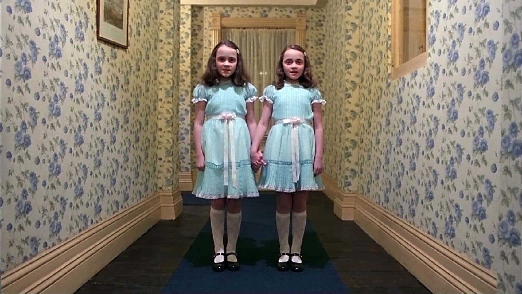 The Grady Twins, wearing light blue dresses with pink bows, stand side by side at the end of a long hallway with blue carpet, and blue floral wallpaper.