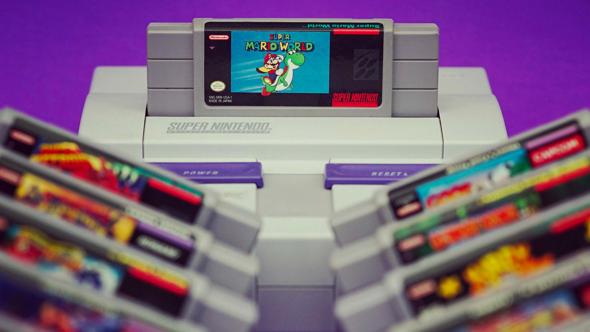 An SNES system sits on a purple backdrop with a variety of blurred out game cartridges infront of it
