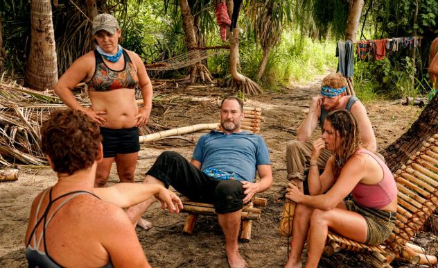 A man in a blue shirt is surrounded by Survivor plays sitting on wooden chairs in a forest
