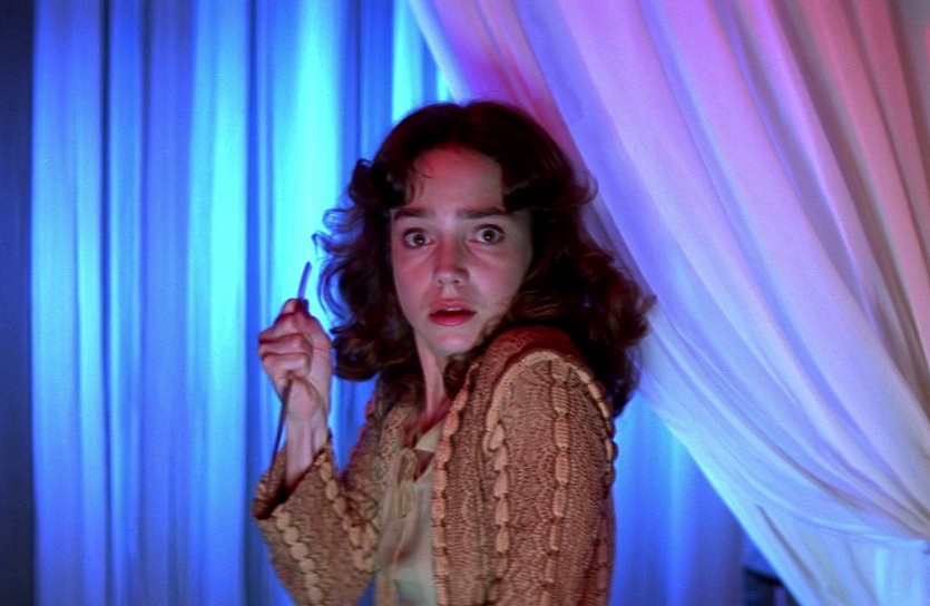 a horrified Suzy stares toward the camera. The room she's in is filled with a collection of white curtains draped from the ceiling. The lighting is a soft pink and blue.