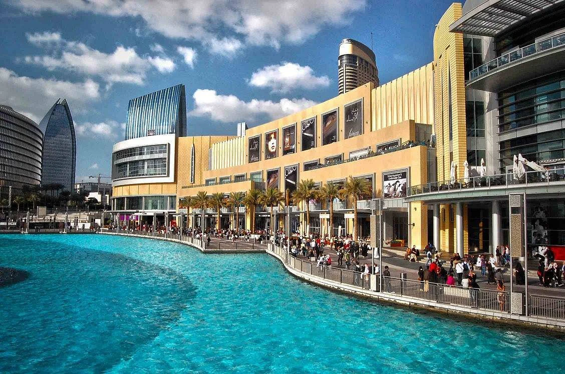 A bustling waterfront promenade outside a modern shopping mall, with a turquoise artificial lake and contemporary buildings in the background.