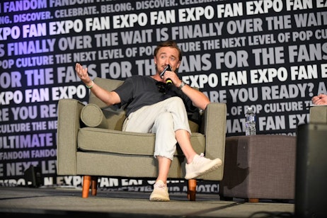 Tom Felton smiles as he tells the audience a story. He sits in the grey wingback chair with his legs crossed and arm extended towards the crowd
